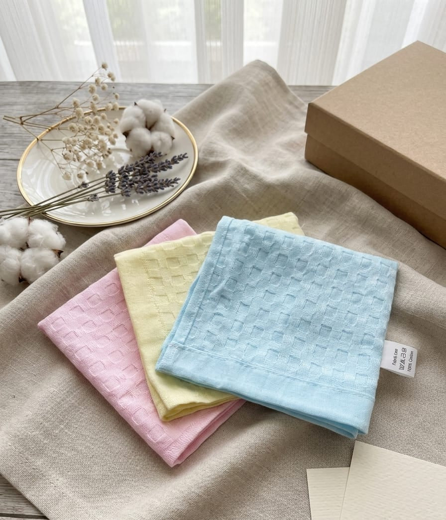 Three textured cotton napkins in pastel pink, pale yellow, and light blue, arranged on a beige linen cloth next to a brown kraft box, with dried flowers and cotton props in the background.